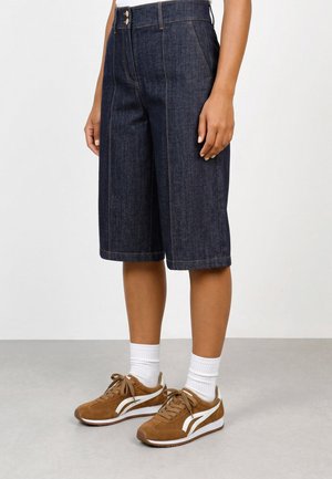 Personne portant un short bermuda en denim bleu foncé taille haute, des chaussettes blanches et des baskets marron avec des rayures blanches, debout sur un sol gris.