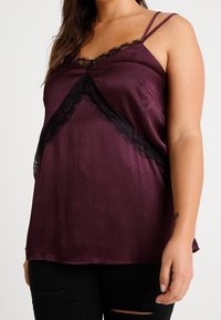 Kvinna klädd i en ärmlös lila satinöverdel med svart spetsdetaljering och tunna axelband, kombinerad med trasiga svarta byxor.