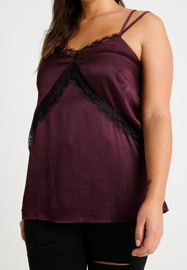 Kvinna klädd i en ärmlös lila satinöverdel med svart spetsdetaljering och tunna axelband, kombinerad med trasiga svarta byxor.