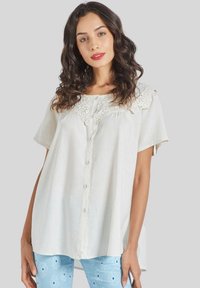 Blusa blanca de botones con mangas cortas, que presenta un corsé de encaje y un ajuste relajado. Combinada con pantalones de flores en tonos azul claro.