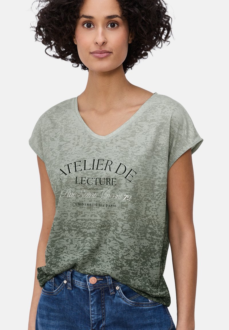 Mujer con cabello rizado que lleva una camiseta verde con degradado y cuello en V con texto en francés y jeans de mezclilla azules, de pie contra un fondo blanco.