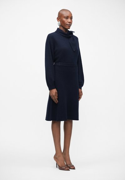 Vestido de malha azul-marinho com gola alta e detalhe de laço, apresentando mangas longas e saia evasê, combinado com sapatos de salto em tom de castanho.