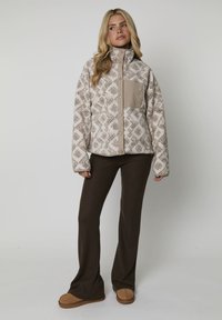 Giacca in pile beige con motivo a diamante, colletto alto e tasca laterale; abbinata a pantaloni svasati marrone scuro e pantofole marroni.