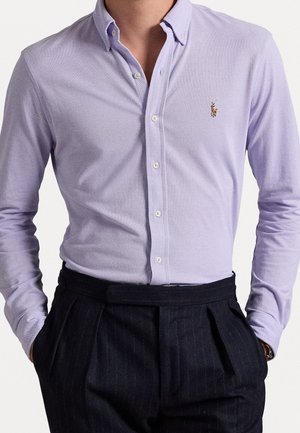 Homme portant une chemise à boutons violet clair avec un petit logo brodé sur la poitrine et un pantalon plissé foncé, les mains dans les poches.