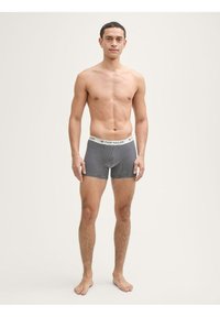 Moški črno-beli prugasti bokserji z mehkimi elastikami, označenimi z "TOM TAILOR", s tesnim prilegajočim fitom. Model stoji bos.