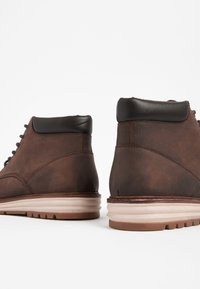 Hnedé kožené členkové čižmy s tmavohnedým polstrovaným golierom, béžovou podrážkou a šnurovaním, s hladkou textúrou a minimalistickým zdobením.