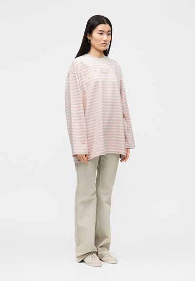 Γυναίκα με μακριά σκούρα μαλλιά που φοράει ένα υπερμεγέθη ροζ και λευκό ριγέ φούτερ, μπεζ παντελόνια φαρδιάς γραμμής και μπεζ παπούτσια, στέκεται μπροστά σε απλό φόντο.