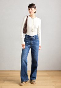 White ribbed long-sleeve shirt with button placket, paired with dark blue high-waisted jeans and brown shoulder bag. Casual footwear.