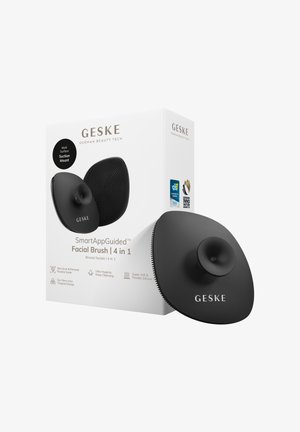 Zwarte ovale siliconen gezichtsborstel met zuignap en een gestructureerd oppervlak, naast witte verpakking met het label "GESKE SmartAppGuided Facial Brush 4 in 1."