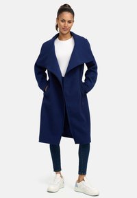 Manteau long bleu marine avec un large col, fabriqué en tissu doux, comportant des poches avant et une fermeture éclair. Porté sur un haut blanc avec un jean foncé.