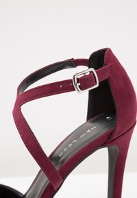 Sandale à talon haut en daim bordeaux avec une large bride, une fine bride de cheville avec une boucle argentée, et une semelle intérieure noire lisse.