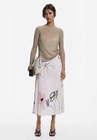 Beiger Strickpullover, pinker Midirock mit Blumenmuster, marineblaue Sandaletten mit Absatz, graue Handtasche, schwarzes Handy in der Hand.