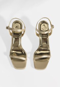 Goldene Riemchen-High-Heelsandalen mit glänzendem Metallic-Finish, quadratischer Zehenpartie und Knöchelriemen, die eine reflektierende, glatte Oberfläche aufweisen.