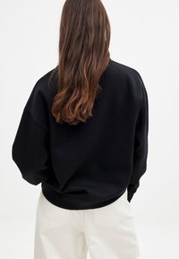 Sudadera negra de gran tamaño con hombros caídos y puños acanalados, combinada con pantalones de color claro. Textura suave, con un detalle minimalista.