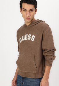 Bruine hoodie van zacht materiaal, met een voorzak, geribde manchetten en het "GUESS" logo in het wit over de borst.