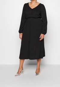 Svart långärmad klänning med V-ringning, åtsittande midja och prickmönster; kombinerad med eleganta silverhöga klackar. Slät tygtextur.