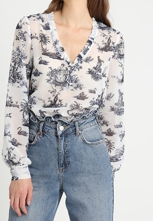 Kvinde iført en tynd, hvid bluse med sort pastoral og svaneprint, kombineret med højtaljede blå jeans med en flæset talje.