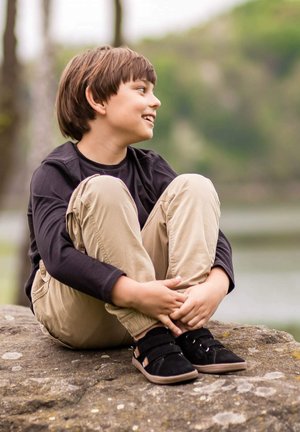 Schwarzes Langarmshirt, beige Hose und schwarze Schuhe mit Klettverschlüssen. Sitzt seitlich auf einem Stein am Wasser. Natürlicher Hintergrund.
