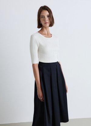 Donna in piedi davanti a uno sfondo semplice, indossa un maglione bianco a coste e una gonna nera plissettata a vita alta.