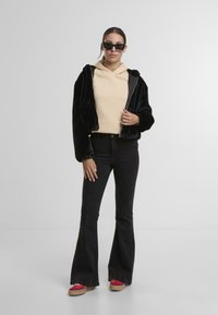 Chaqueta de piel sintética negra con cremallera sobre una sudadera beige, combinada con jeans negros acampanados y zapatos de plataforma rojos. El modelo lleva gafas de sol negras.