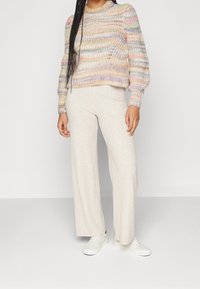 Maglione a maglia con righe pastello e texture a coste, abbinato a pantaloni beige chiaro a gamba larga. Sneakers bianche completano l'outfit.