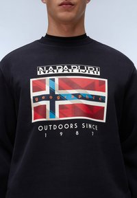 Marine sweatshirt met een geprinte afbeelding van een vlag met rode, blauwe en beige elementen. De tekst luidt "NAPAPIJRI" en "OUTDOORS SINCE 1987."