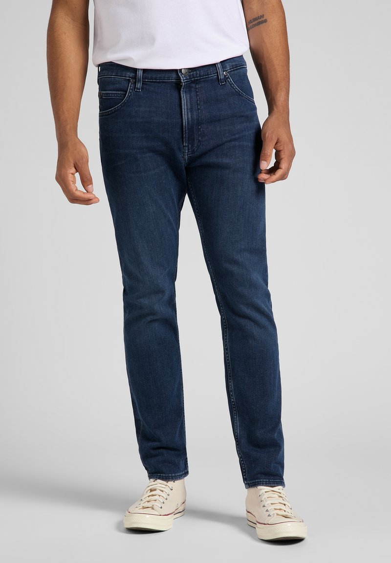 Hombre vistiendo jeans slim-fit de color azul oscuro, zapatillas blancas y una camiseta blanca, de pie contra un fondo gris liso.