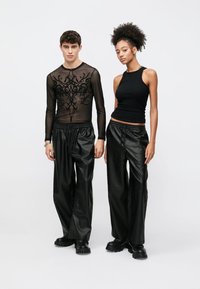 Μαύρο διάφανο μακρυμάνικο τοπ με κεντήματα με λουλούδια, σε συνδυασμό με μαύρο ψηλόμεσο, γυαλιστερό παντελόνι φαρδιάς γραμμής και χοντρά μαύρα παπούτσια.