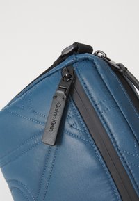 Blauwe gewatteerde nylon tas met een zwarte rits, voorzien van een gestructureerd oppervlak en een Calvin Klein logo-label. Compact ontwerp, geschikt voor dagelijks gebruik.