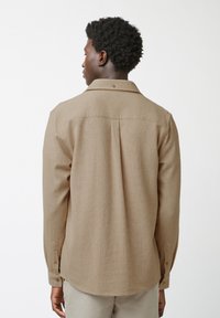 Camicia lunga a maniche di colore marrone, realizzata in tessuto intrecciato, con colletto classico, chiusura con bottoni e un dettaglio di cucitura centrale sul retro.