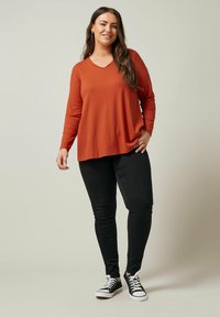Langærmet, ribbet top i rustfarve med v-udskæring, parret med sorte skinny jeans og sorte sneakers med hvide detaljer.