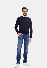 Marineblauw vest met geribde onderrand en manchetten, gedragen over een witte overhemd met kraag, gecombineerd met blauwe jeans en witte sneakers. Gesponnen stof, ontspannen pasvorm.