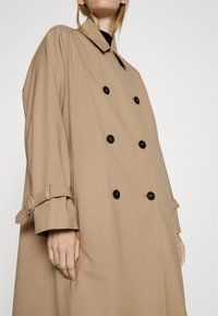Personne portant un trench-coat beige croisé avec des boutons noirs et des détails de ceinture sur les manches, sur un fond clair uni.