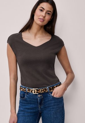 Frau trägt ein dunkelbraunes Top mit Kappärmel, blaue Jeans mit Leopardenmuster-Gürtel, steht mit einer Hand in der Tasche vor einfarbigem Hintergrund.