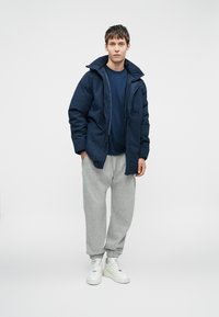 Mornarska podložena jakna z visokim ovratnikom, v kombinaciji s temno modro majico, sivimi trenirkami in belimi supergami. Gladek material in sproščen kroj.