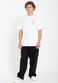 White short-sleeve cotton t-shirt featuring a pink spiral graphic on the left chest. Paired with black wide-leg pants and white sneakers.