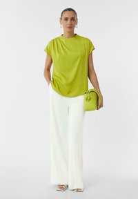 Limonkowa bluzka z krótkim rękawem, luźny krój; w zestawieniu z białymi spodniami o wysokim stanie i szerokich nogawkach oraz małą zieloną torebką z przewiązanymi akcentami.