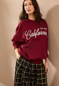 Mørkerød hoodie med hvid "California EST 1989" tryk, parret med en plisseret ternet nederdel i brune og grønlige toner. Blød stofstruktur.
