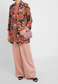 Blazer floreale in verde scuro, rosa e arancione, abbinato a una blusa rosa e pantaloni in satin a gamba larga; tiene in mano una piccola borsa lavanda.