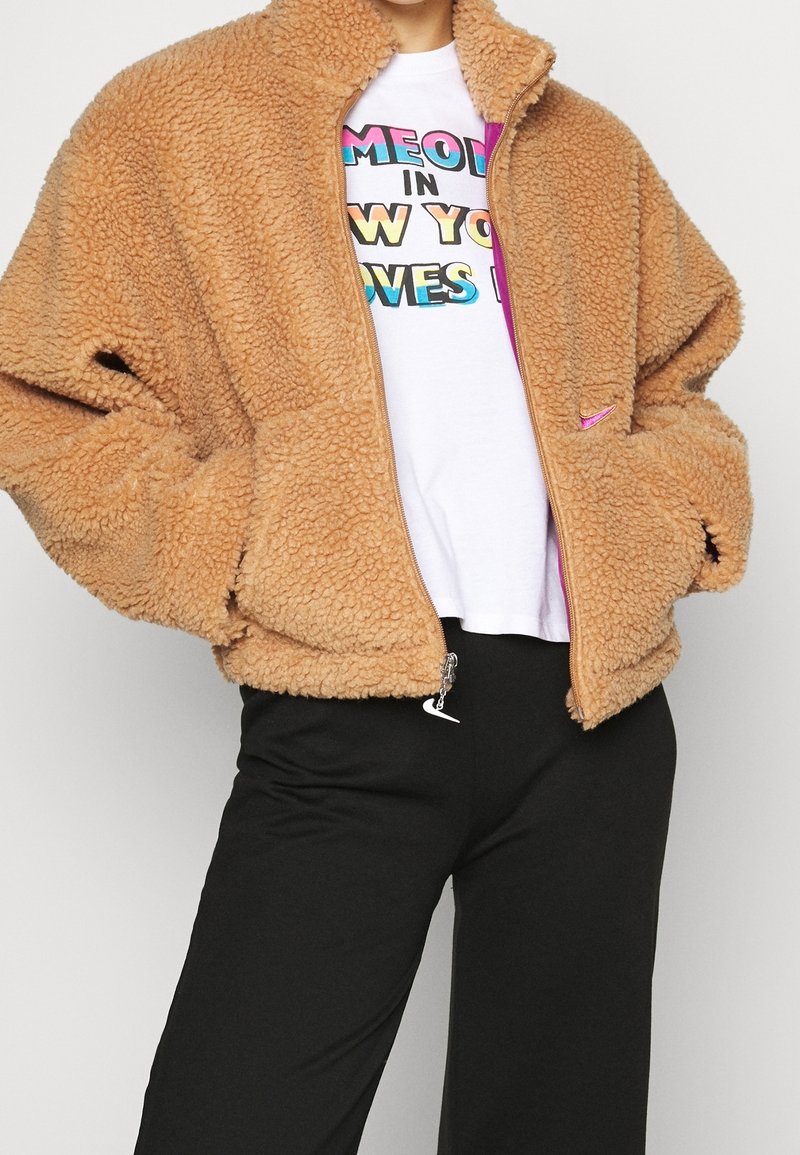 Beige fleecejacka med hög krage, dragkedja och sidofickor. Matchad med en vit t-shirt med färggd text och svarta byxor.