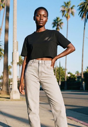 Jeune femme noire portant un t-shirt noir court et un jean clair taille haute, debout sur un trottoir ensoleillé avec des palmiers en arrière-plan.