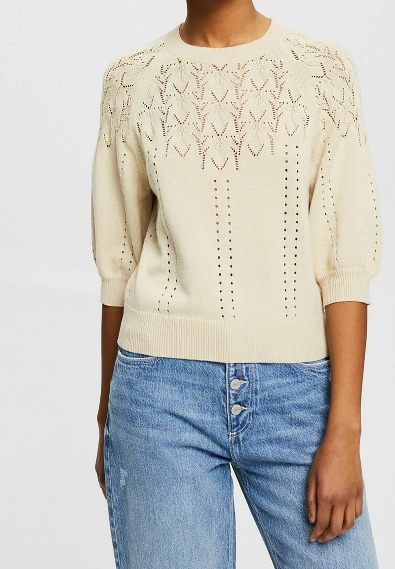 Pull en maille crème avec des motifs de feuilles ajourées et des manches trois-quarts, associé à un jean bleu taille haute avec trois boutons.
