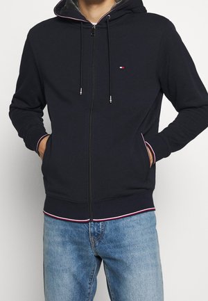 Mand med hænderne i lommerne iført sort lynlåshoodie med lille logo og lyseblå jeans mod hvid baggrund.