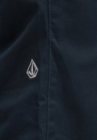 Το σκουρόχρωμο ναυτικό ύφασμα διαθέτει κεντημένο ανοιχτό γκρι λογότυπο που μοιάζει με στα γόνα, τοποθετημένο στην αριστερή πλευρά, με ομαλή υφή.