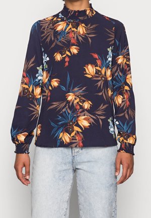 Person iført en navyblå langærmet bluse med orange og blå blomstermønster, smockede manchetter og halsudskæring, kombineret med lyseblå jeans.