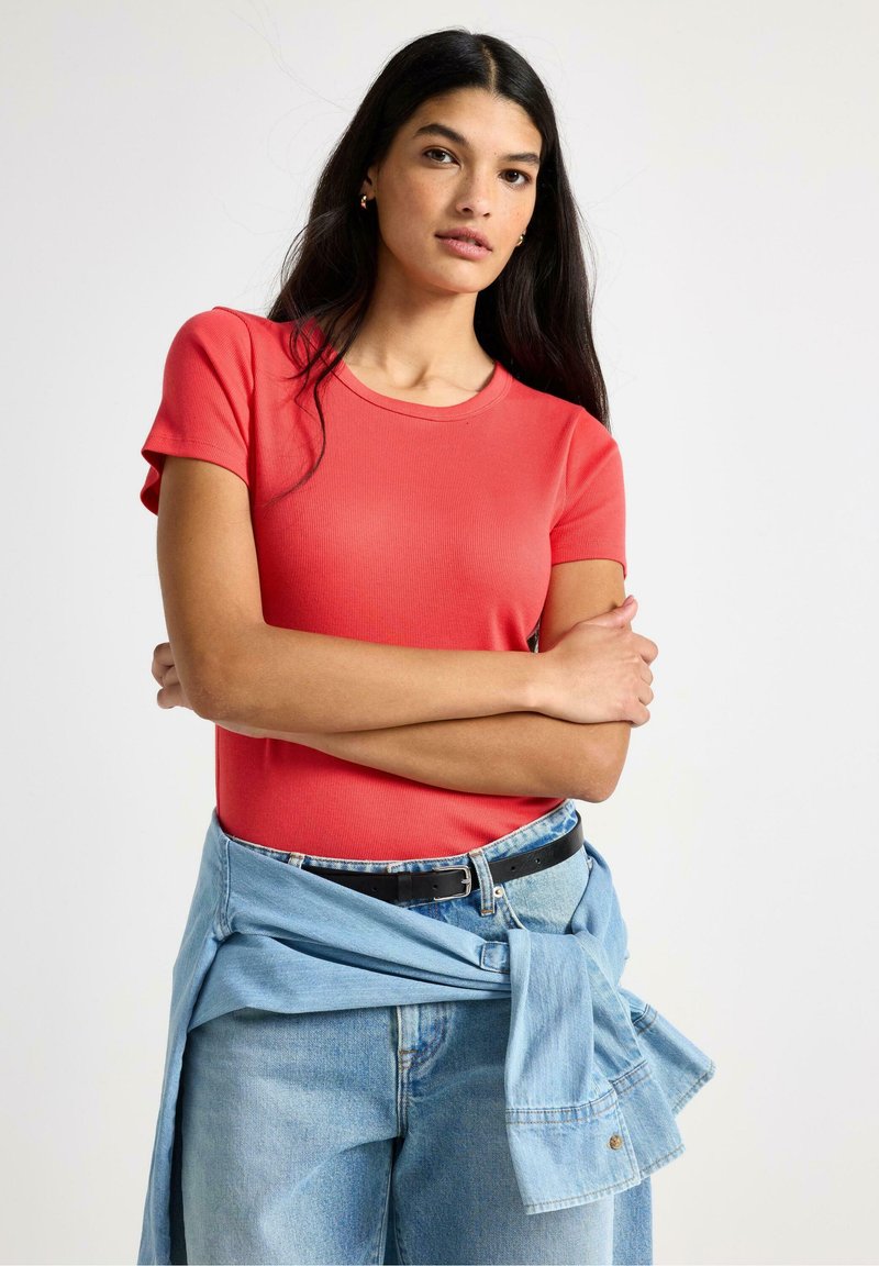 Rød ribbet kortærmet t-shirt kombineret med lyseblå denimjeans og en denimjakke bundet om livet. Modellen har langt, mørkt hår.