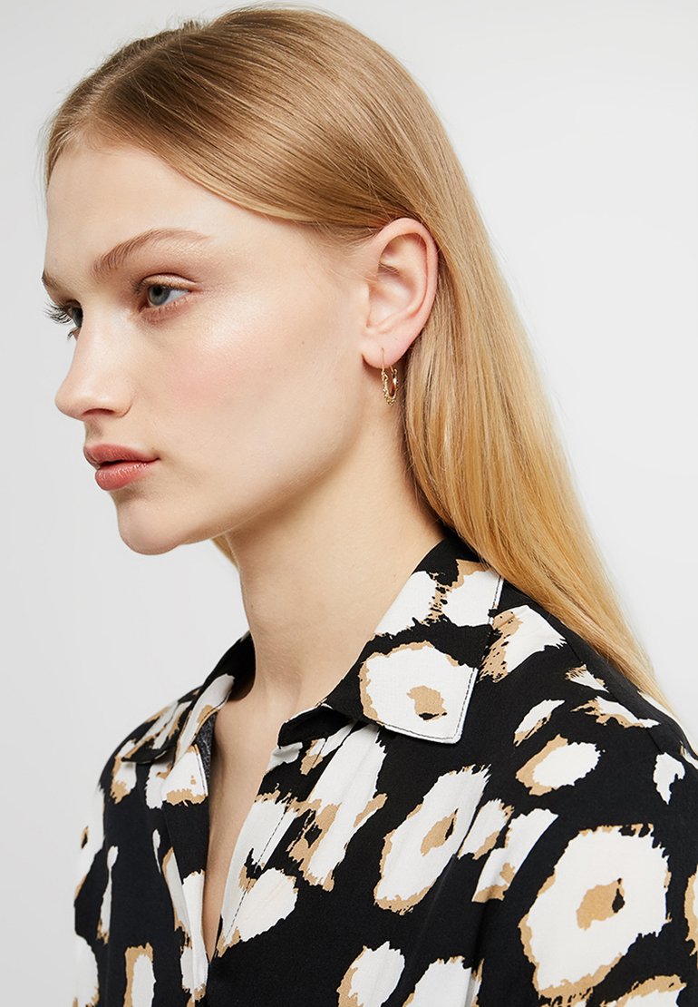 Svart skjorta med ett iögonfallande mönster av vita och bruna blommor, med krage och knäppning. Modellen bär små hoopörhängen; rakt blont hår.