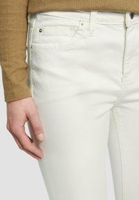 Witte denim jeans hebben een klassieke vijf-pocket ontwerp met zichtbare stiksels en metalen accenten, gecombineerd met een getextureerde bruine top.