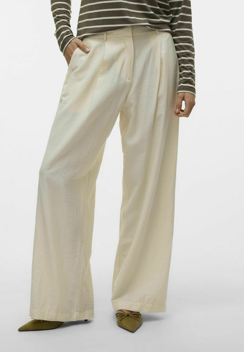 Pantalons évasés en tissu crème clair, avec des plis sur le devant et des poches latérales, associés à des talons aiguilles verts olive. Haut rayé visible.