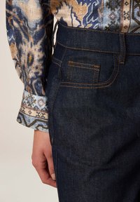 Los jeans de mezclilla oscura presentan detalles de costura en color naranja, con un bolsillo lateral y una cinturilla ajustada. La camisa estampada tiene tonos azules y beige.